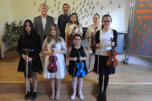 Grupa uczniów ze skrzypcami stoi na scenie z nauczycielami